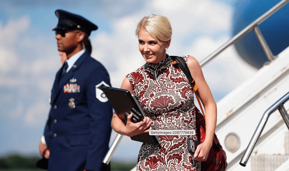 A woman in a patterned dress walks down the steps of an aircraft, holding a laptop and a bag. In the background, a uniformed military officer stands guard.