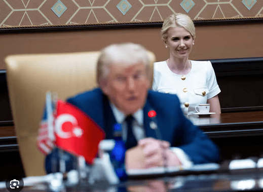 A blurred view of a man in a suit, possibly engaged in a discussion, with two flags in the foreground, one Turkish and one American. In the background, a woman with light hair in a white dress smiles while seated at a table.