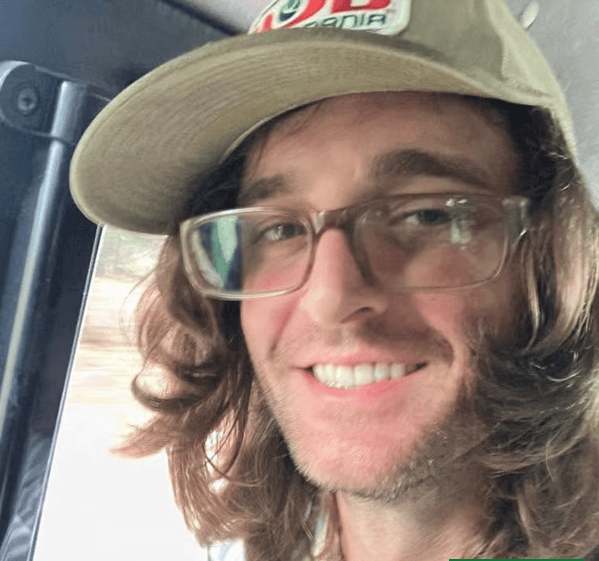 A smiling individual with long hair wearing a cap and glasses, sitting inside a vehicle.