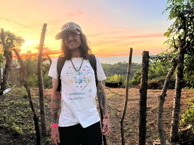 A person standing outdoors with a sunset backdrop, wearing a white t-shirt with colorful graphics, sunglasses, and a cap, surrounded by wooden posts and greenery.